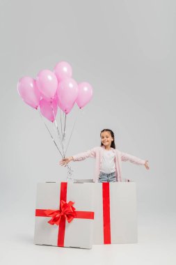 Carefree preteen kid in casual clothes looking at camera while holding pink festive balloons while standing in big present during international children day celebration on grey background clipart