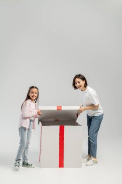 Gri arka planda uluslararası çocuklar gününde büyük hediye kutusunu açarken gündelik kıyafetlerini giymiş neşeli anne ve reşit olmayan kız çocukları kameraya bakıyor.