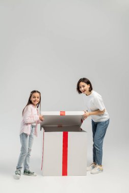 Full length of cheerful mother and preteen daughter in casual clothes looking at camera while opening big present box during international children day on grey background clipart