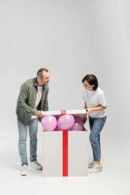 Full length of smiling adult couple in casual clothes opening big gift box with pink balloons during child protection day celebration while standing on grey background clipart