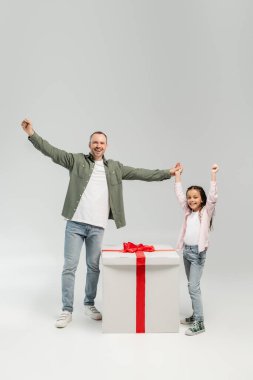 Heyecanlı ve kaygısız baba ve reşit olmayan kızın, gri arka planda çocuk koruma günü kutlamalarında büyük bir hediyeye yakın kameraya bakması.