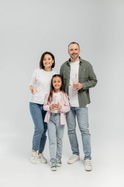 Full length of smiling family with preteen daughter in casual clothes holding milkshakes in plastic cups and looking at camera during children day celebration on grey background clipart