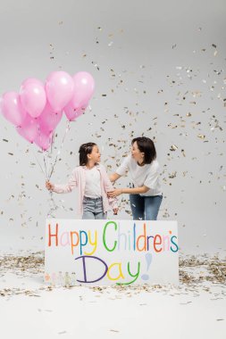 Side view of cheerful woman in casual clothes hugging preteen daughter with pink balloons while standing together near placard with happy children day under falling confetti on grey background clipart