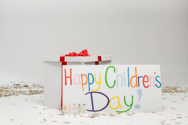 Renkli mutlu çocuk günü yazılı pankart. Büyük hediye kutusunun yanında. Arka planda gri bir fiyonk ve şenlikli konfeti var. 
