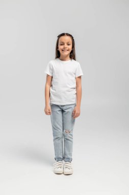 Beyaz tişörtlü ve kot pantolonlu neşeli ve reşit olmayan bir kız küresel çocuk koruma gününü kutlarken kameraya bakıyor ve gri arka planda duruyor.