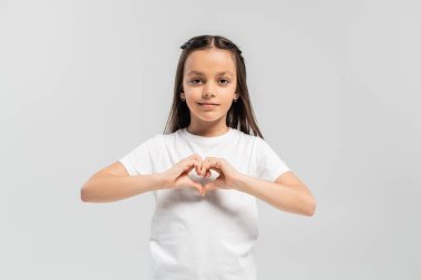 happy preteen girl with long brunette hair standing in white t-shirt and showing heart gesture with hands isolated on grey background, child protection day holiday  clipart