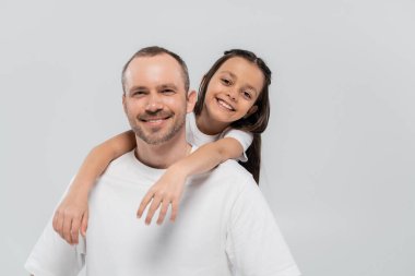 happy preteen girl with long brunette hair leaning on unshaved and cheerful father with bristle while posing in white t-shirts and looking at camera on grey background, Child protection day  clipart