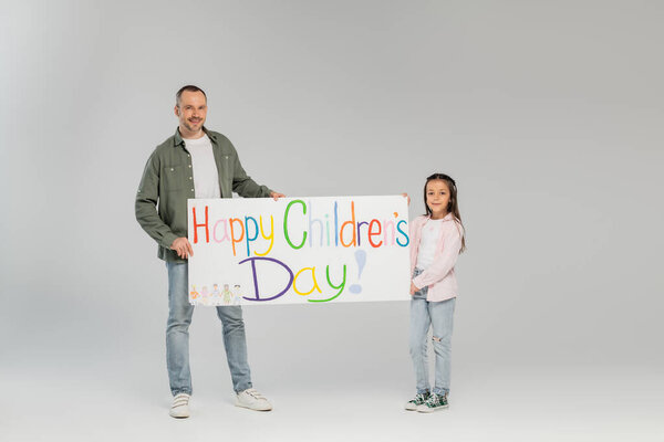 Full length of smiling father and preteen daughter in casual clothes looking at camera while holding placard with happy children's day lettering while celebrating together on grey background