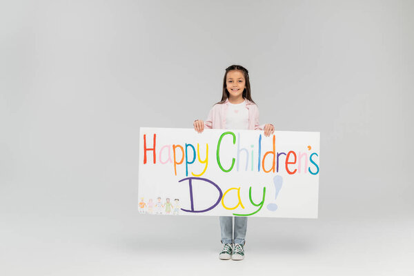 Full length of cheerful preteen girl in casual clothes looking at camera while holding placard with happy children's day lettering while standing on grey background with copy space