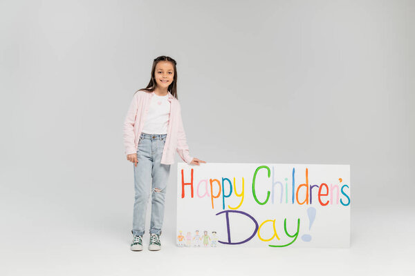 Smiling preteen girl in casual clothes looking at camera while standing near placard with happy children's day lettering during holiday in June on grey background with copy space 