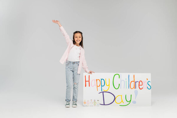 Full length of cheerful preteen girl in casual clothes waving hand and looking at camera near placard with happy children's day lettering and standing on grey background
