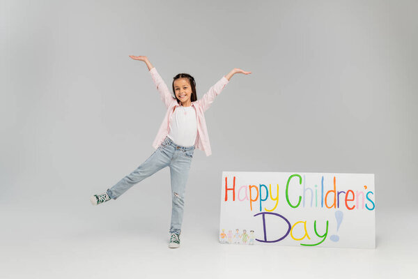 Excited and positive preteen girl in casual clothes having fun and looking at camera near placard with happy children's day lettering during celebration on grey background