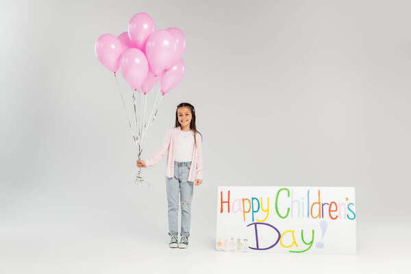 Full length of smiling preteen kid in casual clothes holding pink balloons and looking at camera near placard with happy children's day lettering during celebration on grey background