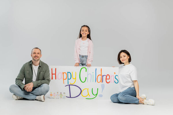 Full length of preteen kid in casual clothes looking at camera while standing near smiling parents and placard with happy children's day lettering during holiday on grey background