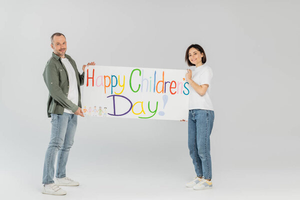 Full length of smiling adult couple in casual clothes looking at camera while holding placard with happy children's day lettering together while standing on grey background