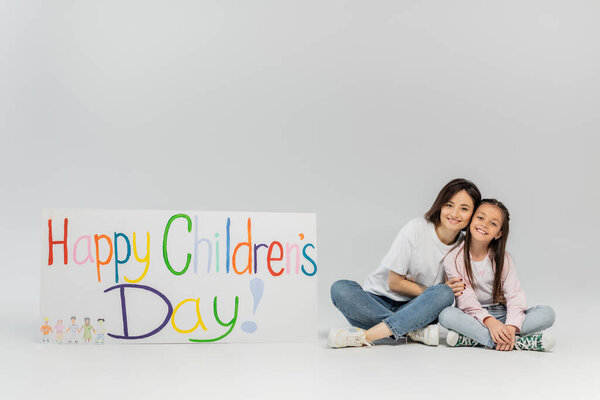 Full length of smiling mother in casual clothes hugging preteen daughter and looking at camera together near placard with happy children's day lettering on grey background