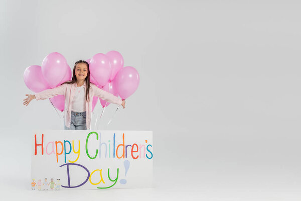 Cheerful and preteen girl in casual clothes looking at camera near pink festive balloons and placard with happy children's day lettering while celebrating on grey background