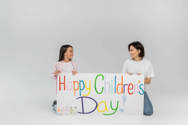 stock image Smiling preteen girl looking at brunette and positive mother near placard with happy children's day lettering during celebration in June on grey background with copy space 