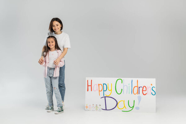 Adult woman in casual clothes hugging cheerful preteen daughter and looking at camera near placard with happy children's day lettering during celebration on grey background
