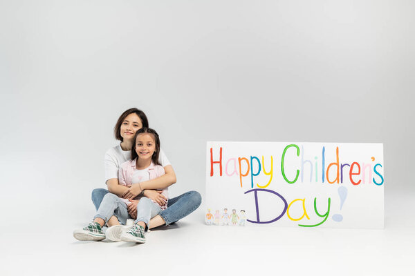 Brunette woman in casual clothes hugging carefree preteen daughter and looking at camera while sitting together near placard with happy children's day lettering on grey background