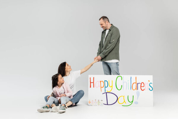 Smiling man holding hand of wife near preteen daughter and placard with happy children's day lettering during kid protection day celebration on grey background