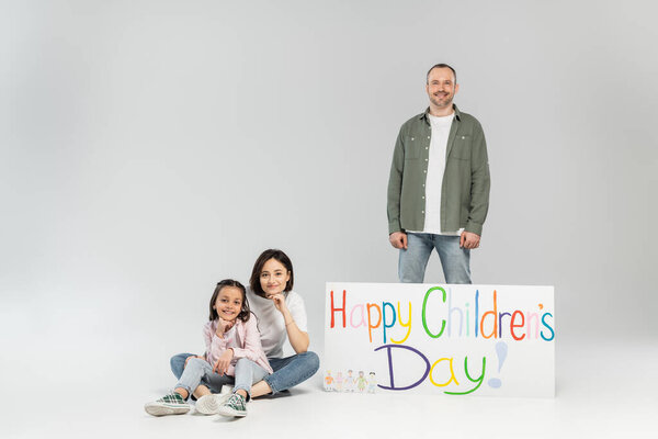 Smiling man looking at camera while standing near wife, preteen daughter and placard with happy children's day lettering during celebration on grey background
