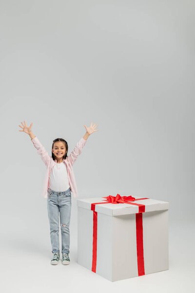 Carefree preteen girl in casual clothes waving hands at camera while standing near big gift box with bow during international children day celebration on grey background