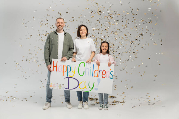 Full length of cheerful family with preteen daughter in casual clothes holding placard with happy children's day lettering and standing under falling festive confetti on grey background