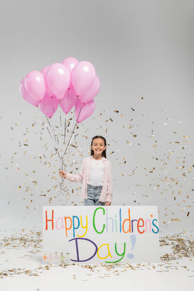 Cheerful preteen kid in casual clothes looking at camera while holding pink balloons near placard with happy children's day lettering and under falling festive confetti on grey background