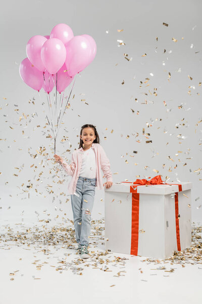 Full length of carefree preteen girl in casual clothes holding pink balloons and looking at camera near big gift box while standing under confetti during child protection day celebration on grey background
