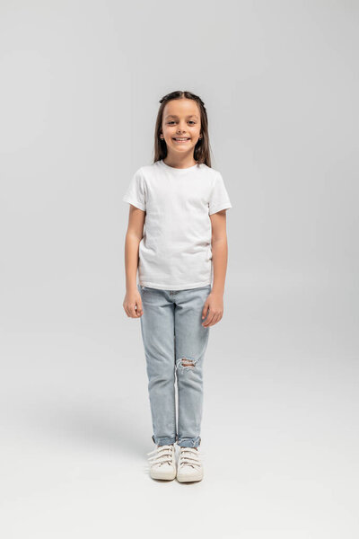 Full length of cheerful and preteen girl in white t-shirt and jeans looking at camera while celebrating global child protection day and standing on grey background