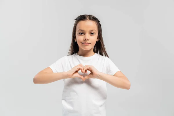 stock image happy preteen girl with long brunette hair standing in white t-shirt and showing heart gesture with hands isolated on grey background, child protection day holiday 