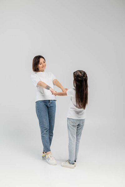 full length of happy mother with tattoo on hand and short hair holding hands with preteen daughter while standing together in white t-shirts and blue denim jeans on grey background 
