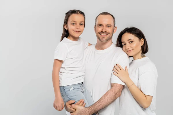 portrait of happy family in white t-shirts looking at camera on grey background, Child protection day, cheerful father lifting preteen daughter near wife with short hair and tattoo