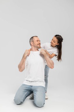 happy preteen girl with long brunette hair leaning on unshaved and cheerful father with bristle while posing in white t-shirts and denim jeans while looking at each other on grey background, Child protection day  clipart