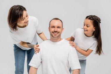 Kısa saçlı, elinde dövmesi olan bir kadın ve kot pantolonlu esmer bir kız gri arka planda beyaz tişörtlü, mutlu bir aileye bakıyor. 