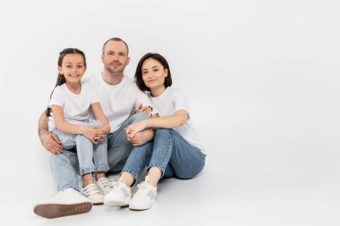 Beyaz tişörtlü ve kot pantolonlu mutlu bir aile kameraya bakıyor ve gri arka planda birlikte oturuyorlar. Uluslararası çocuk koruma günü, ebeveynler ve kızlar. 