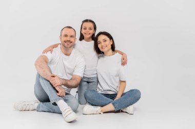 Beyaz tişörtlü ve kot pantolonlu şık bir aile kameraya bakıyor ve gri arka planda birlikte oturuyorlar, uluslararası çocuk koruma günü, reşit olmayan kızın mutlu ebeveynlere sarılması. 