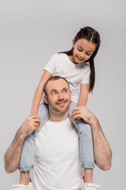 happy preteen daughter with long brunette hair sitting on shoulders of unshaved and cheerful father with bristle on grey background, Child protection day, father and daughter  clipart
