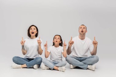 Beyaz tişörtler ve mavi kot pantolonlar içinde oturan ve parmaklarıyla işaret ederek gri arka plana bakan, baba, dövme yaptırmış anne ve kız çocuğuna bakan hayret dolu bir aile. 