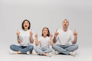 amazed family sitting with crossed legs in white t-shirts and blue denim jeans while pointing with fingers and looking up on grey background, father, tattooed mother and preteen daughter  clipart