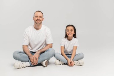 happy preteen girl with long brunette hair and cheerful dad with bristle sitting with crossed legs while posing in white t-shirts and blue denim jeans on grey background, Happy Father`s Day clipart