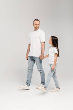Tüylü neşeli bir baba ve beyaz tişörtlü küçük kızı ve kot pantolonu olan ve gri arka planda birlikte yürüyen mutlu babalar günü.