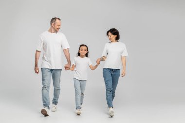 Beyaz tişörtlü, mavi kot pantolonlu, gri arka planda, Haziran 'da uluslararası çocuk koruma gününde el ele tutuşan mutlu aile ve çocukların tam uzunluğunda. 