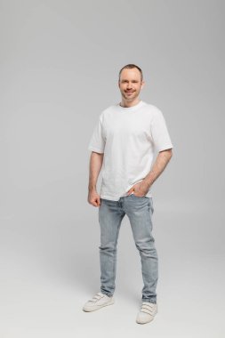 full length of unshaved happy man with bristle standing in white t-shirt and posing with hand in pocket of blue denim jeans while looking at camera on grey background in studio, happiness  clipart