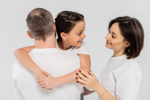 back of father holding happy preteen daughter near cheerful wife with tattoo and short hair smiling while standing in white t-shirts together on grey background, International Day for Protection of Children, 