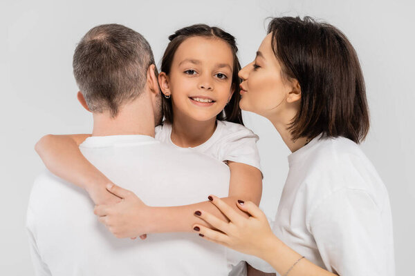 mother with tattoo and short hair kissing cheek of smiling preteen daughter near husband while standing in white t-shirts together on grey background, International Day for Protection of Children, 