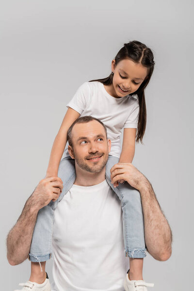 happy preteen daughter with long brunette hair sitting on shoulders of unshaved and cheerful father with bristle on grey background, Child protection day, father and daughter 