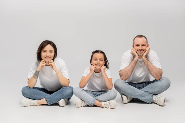 joyous father and tattooed mother with short hair and cheerful preteen daughter sitting with crossed legs in white t-shirts and blue denim jeans on grey background, Happy children's day 
