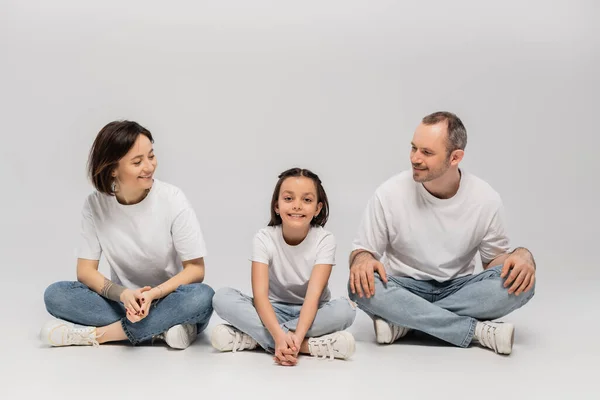 快乐的父亲和纹身的母亲 看着快乐的未成年女儿 双腿交叉 身穿白色T恤衫和灰色背景的蓝色斜纹棉布牛仔裤坐在一起 过着快乐的儿童节 — 图库照片
